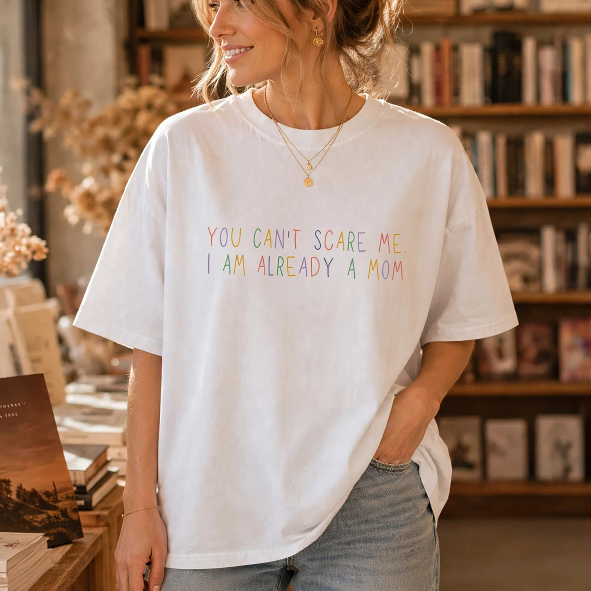 A woman wearing a white graphic t-shirt with the text 'YOU CAN'T SCARE ME I AM ALREADY A MOM' in pastel rainbow colors, standing in a cozy book-filled bookstore.